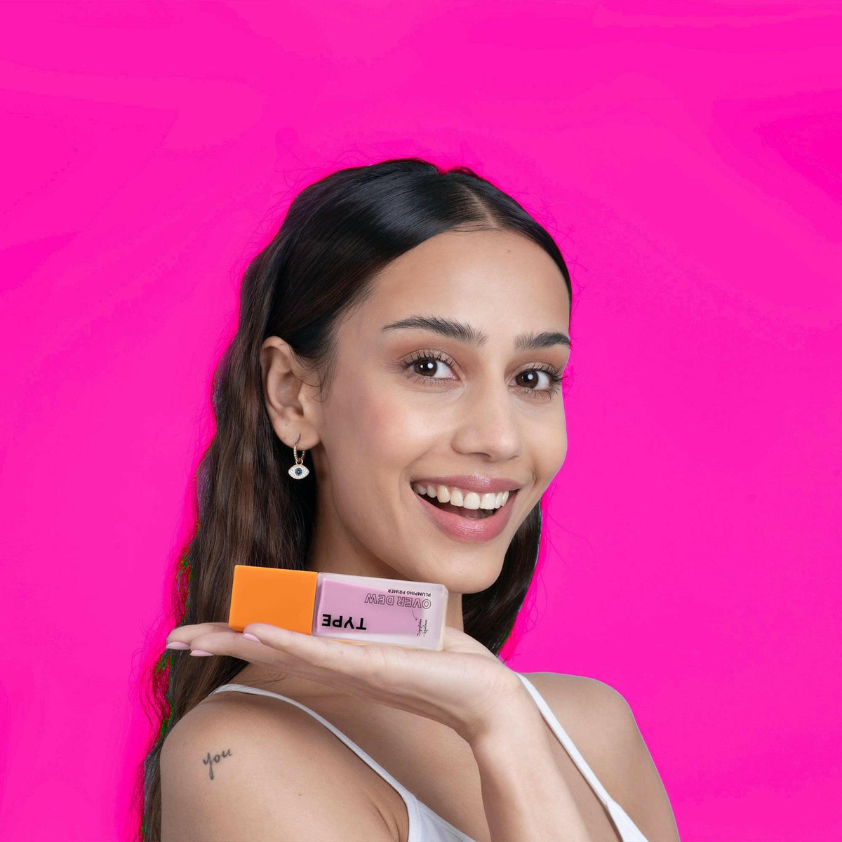 Smiling woman holding OVER DEW PLUMPING PRIMER on bright pink background.